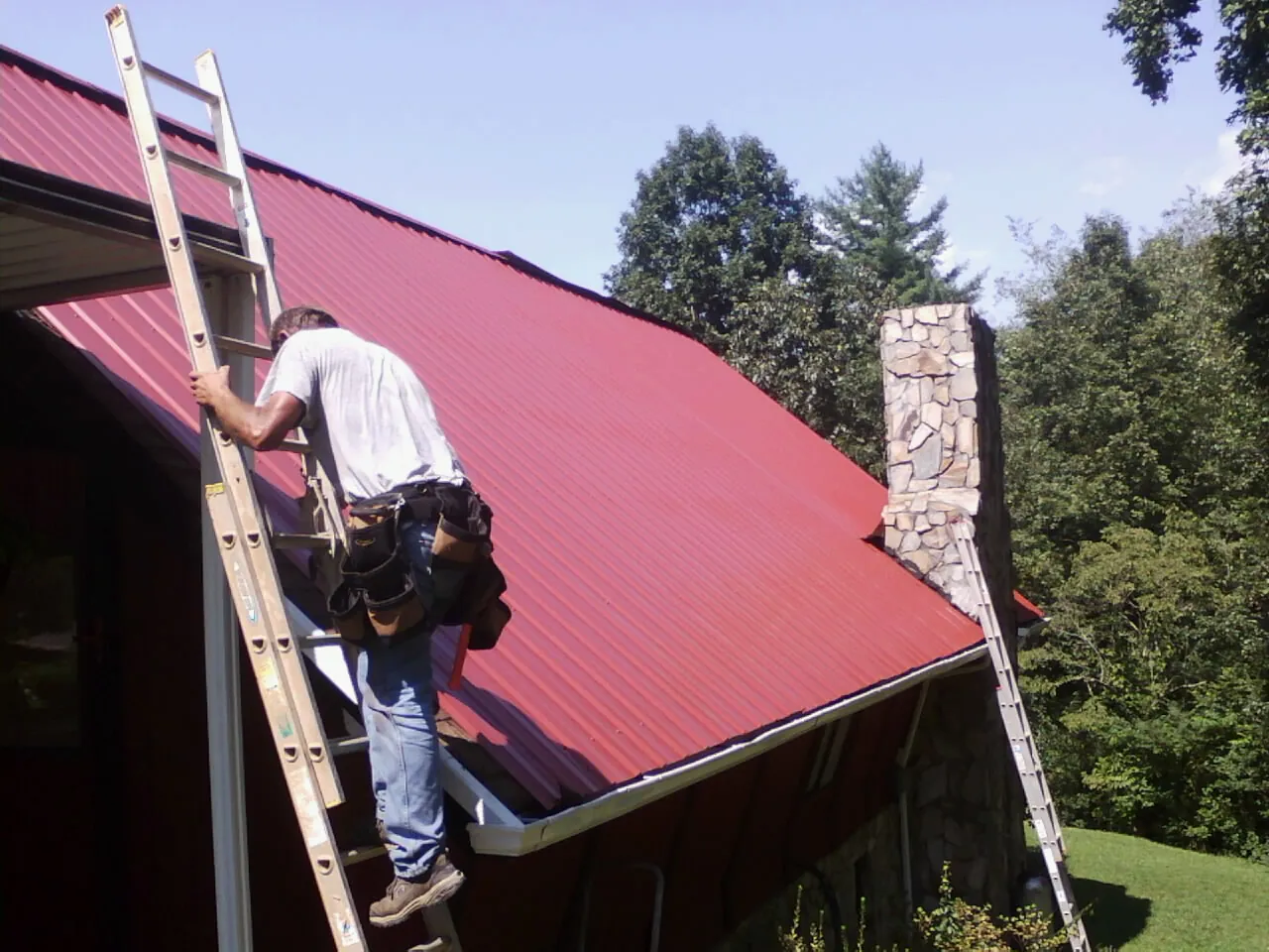 Roofer on a ladder beside a red metal roof and stone chimney — Soffit & Fascia Repair by Roofing Brothers in Frankfort